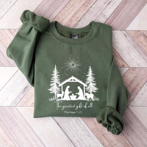 May include: An olive green sweatshirt featuring a white graphic of a nativity scene with the text "The greatest gift of all" and "Matthew 1:21". The design includes a star, trees, and figures within a stable.
