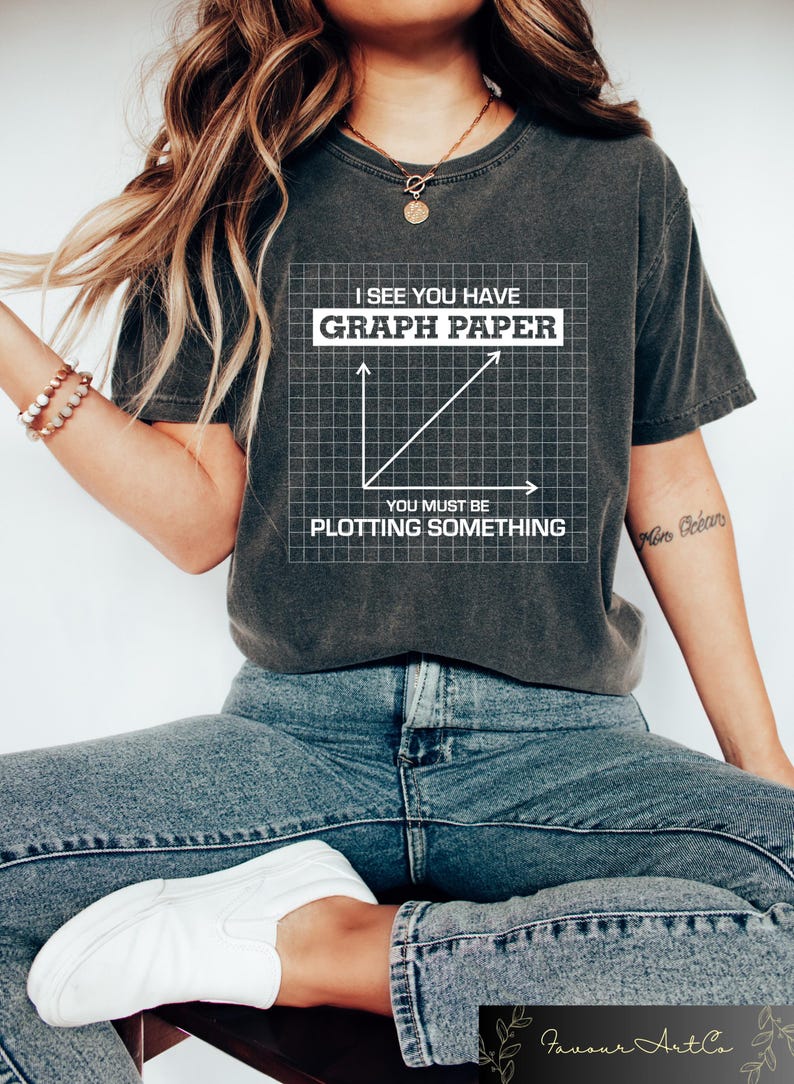 May include: Dark gray t-shirt with the text "I SEE YOU HAVE GRAPH PAPER YOU MUST BE PLOTTING SOMETHING" in white. The graphic includes a graph paper design with a line graph. The shirt is worn with blue jeans and white sneakers.