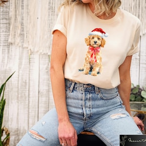 May include: Cream-colored t-shirt featuring a watercolor illustration of a golden doodle puppy wearing a Santa hat and a red bow. The puppy is sitting, and the shirt is paired with blue jeans. The image is set against a neutral background.