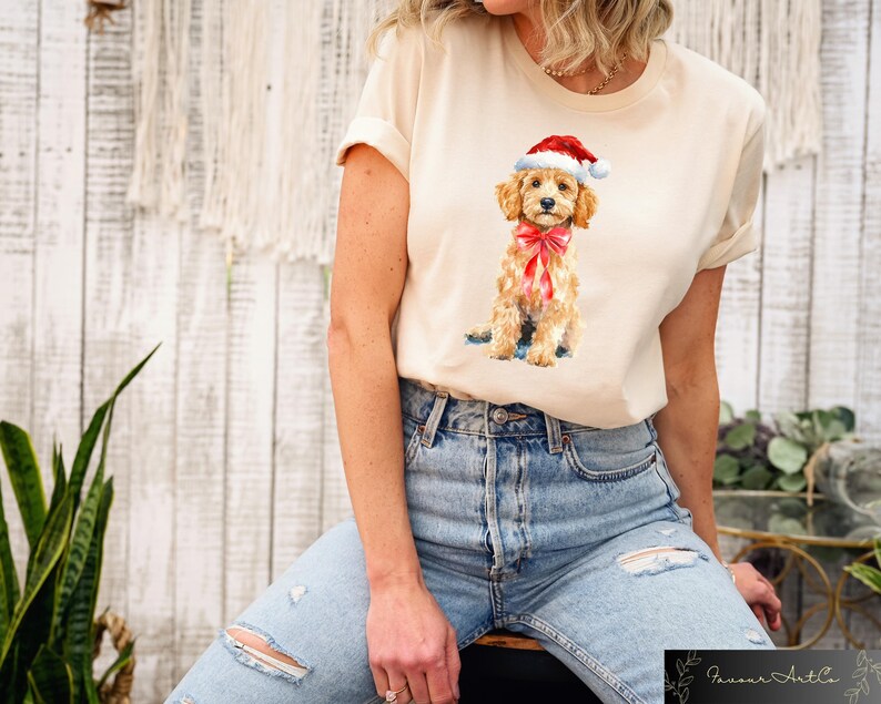 May include: Cream-colored t-shirt featuring a watercolor illustration of a golden doodle puppy wearing a Santa hat and a red bow. The puppy is sitting, and the shirt is paired with blue jeans. The image is set against a neutral background.
