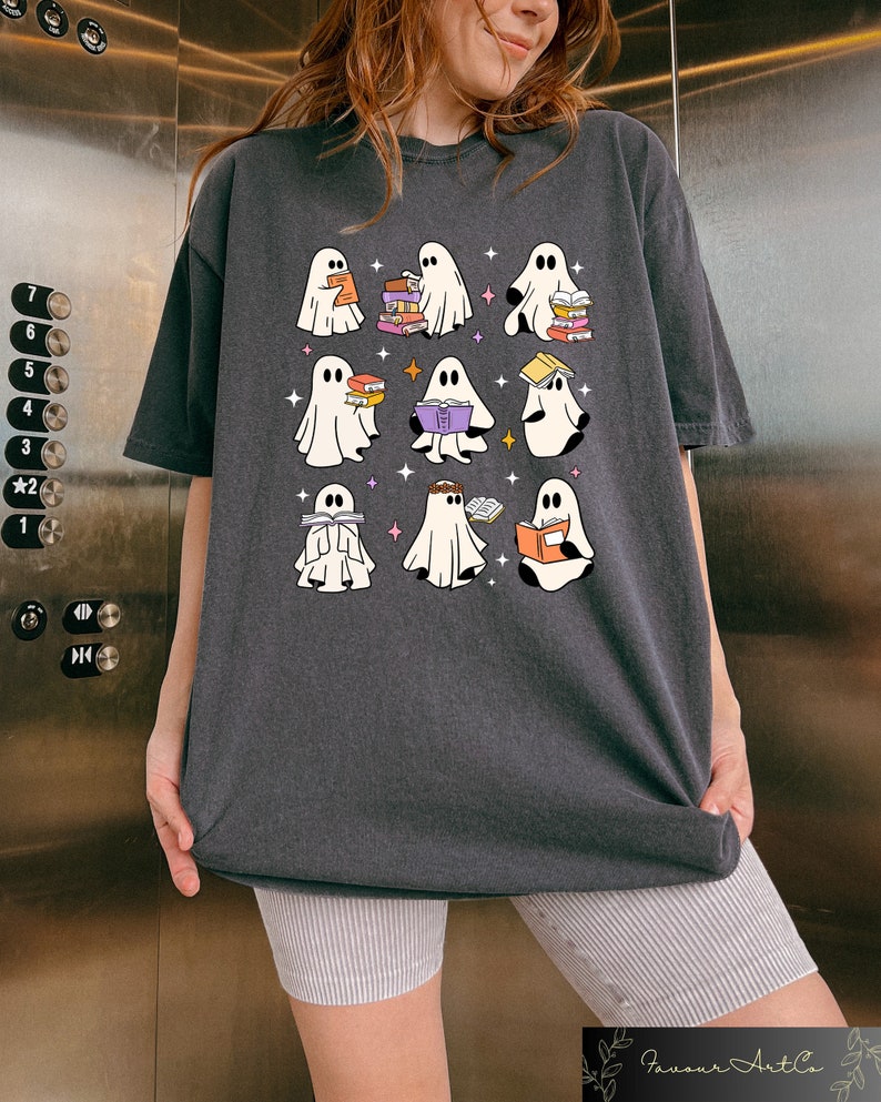 May include: Dark gray t-shirt featuring a whimsical design of nine cartoon ghosts, each holding books. The ghosts are white with black eyes and are surrounded by small stars. The shirt is a comfortable, oversized fit.