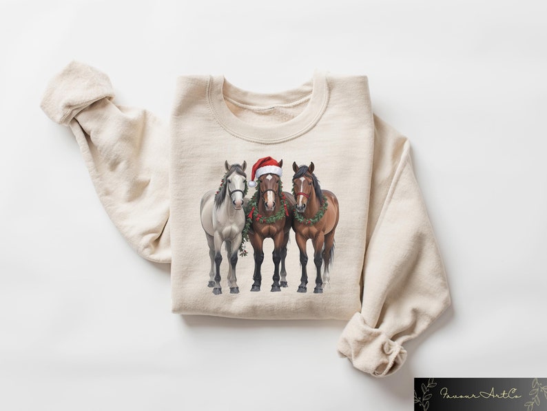 May include: A beige sweatshirt with a Christmas-themed design featuring three horses wearing holly wreaths and a Santa hat. The horses are brown, white, and brown.