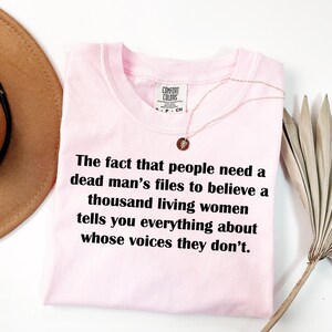 May include: A light pink t-shirt with the text "The fact that people need a dead man's files to believe a thousand living women tells you everything about whose voices they don't." A gold necklace is draped over the shirt.