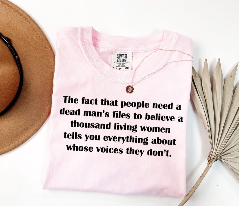 May include: A light pink t-shirt with the text "The fact that people need a dead man's files to believe a thousand living women tells you everything about whose voices they don't." A gold necklace is draped over the shirt.