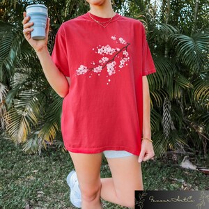Puede incluir: Camiseta roja con una rama de flores de cerezo rosas estampada. La camiseta de manga corta tiene un corte holgado. La persona de la imagen sostiene una taza azul plegable. La camiseta forma parte de la colecci&oacute;n Favour Art Co.