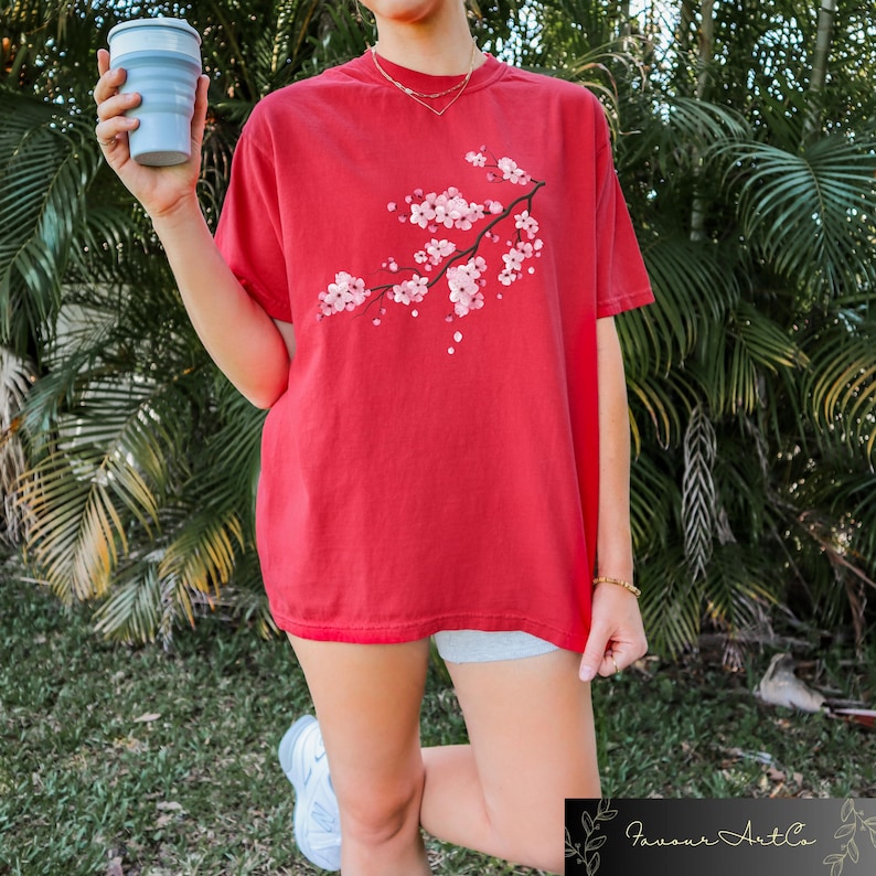 Puede incluir: Camiseta roja con una rama de flores de cerezo rosas estampada. La camiseta de manga corta tiene un corte holgado. La persona de la imagen sostiene una taza azul plegable. La camiseta forma parte de la colecci&oacute;n Favour Art Co.