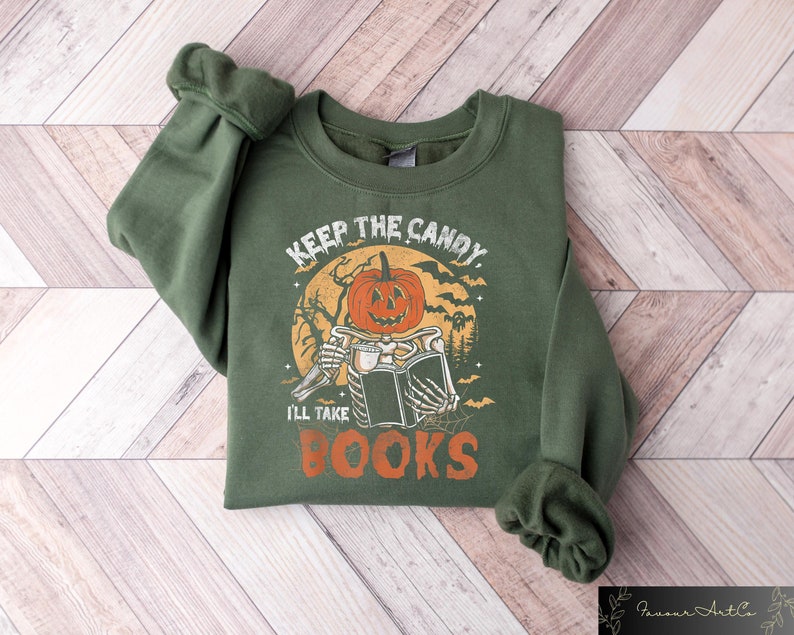 Op de afbeelding: Groen sweatshirt met een Halloween-thema afbeelding. De afbeelding toont een skelet dat een boek en een pompoen vasthoudt met de tekst "Keep the Candy I'll Take Books".
