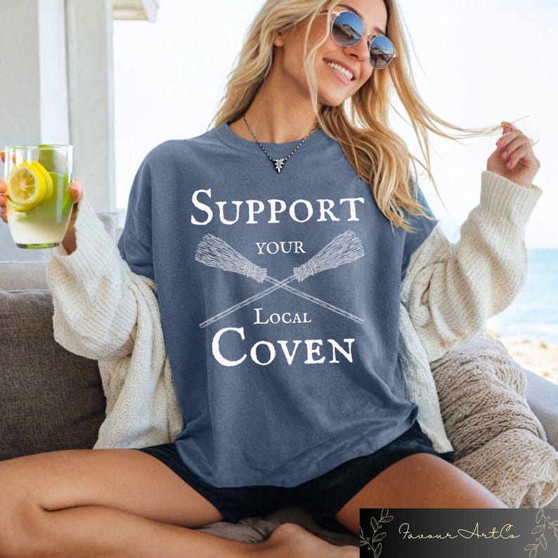 May include: A blue t-shirt with the text "Support Your Local Coven" in white lettering. The text is surrounded by two crossed broomsticks.