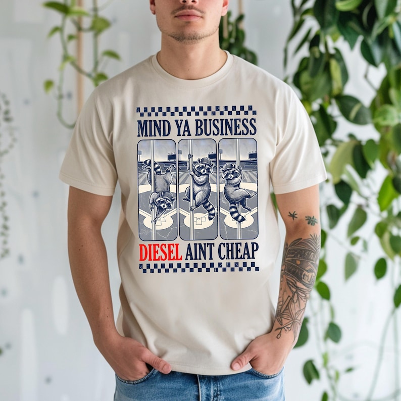 May include: Beige t-shirt featuring a graphic with three raccoons on a baseball field. The text "MIND YA BUSINESS" is above the raccoons, and "DIESEL AINT CHEAP" is below. The raccoons are in various poses.