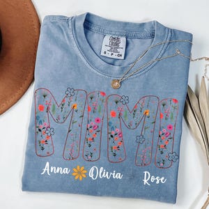 Pode incluir: Uma camiseta azul clara com a palavra "MAMI" em um design floral. Os nomes "Anna", "Olivia" e "Rose" s&atilde;o impressos abaixo. Um colar de ouro est&aacute; drapeado sobre a camiseta.