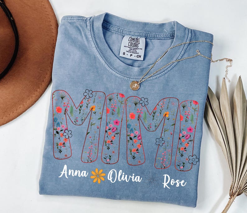 Pode incluir: Uma camiseta azul clara com a palavra "MAMI" em um design floral. Os nomes "Anna", "Olivia" e "Rose" s&atilde;o impressos abaixo. Um colar de ouro est&aacute; drapeado sobre a camiseta.