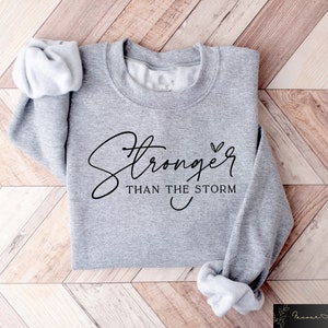May include: Gray sweatshirt with the text "Stronger than the storm" in black cursive lettering with a small heart symbol.
