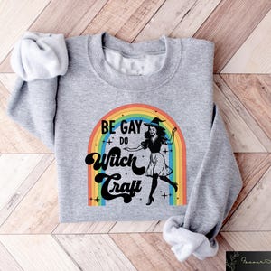 Pode incluir: Um moletom cinza com o texto "BE GAY DO Witch Craft" acima de um arco-&iacute;ris e uma ilustra&ccedil;&atilde;o de bruxa. A bruxa est&aacute; usando um chap&eacute;u e um vestido pretos. O moletom est&aacute; dobrado em uma superf&iacute;cie de madeira.