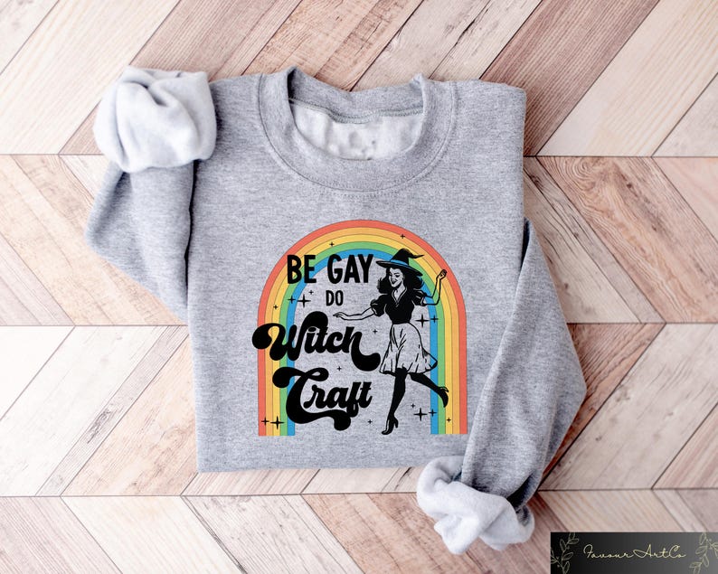 Pode incluir: Um moletom cinza com o texto "BE GAY DO Witch Craft" acima de um arco-&iacute;ris e uma ilustra&ccedil;&atilde;o de bruxa. A bruxa est&aacute; usando um chap&eacute;u e um vestido pretos. O moletom est&aacute; dobrado em uma superf&iacute;cie de madeira.