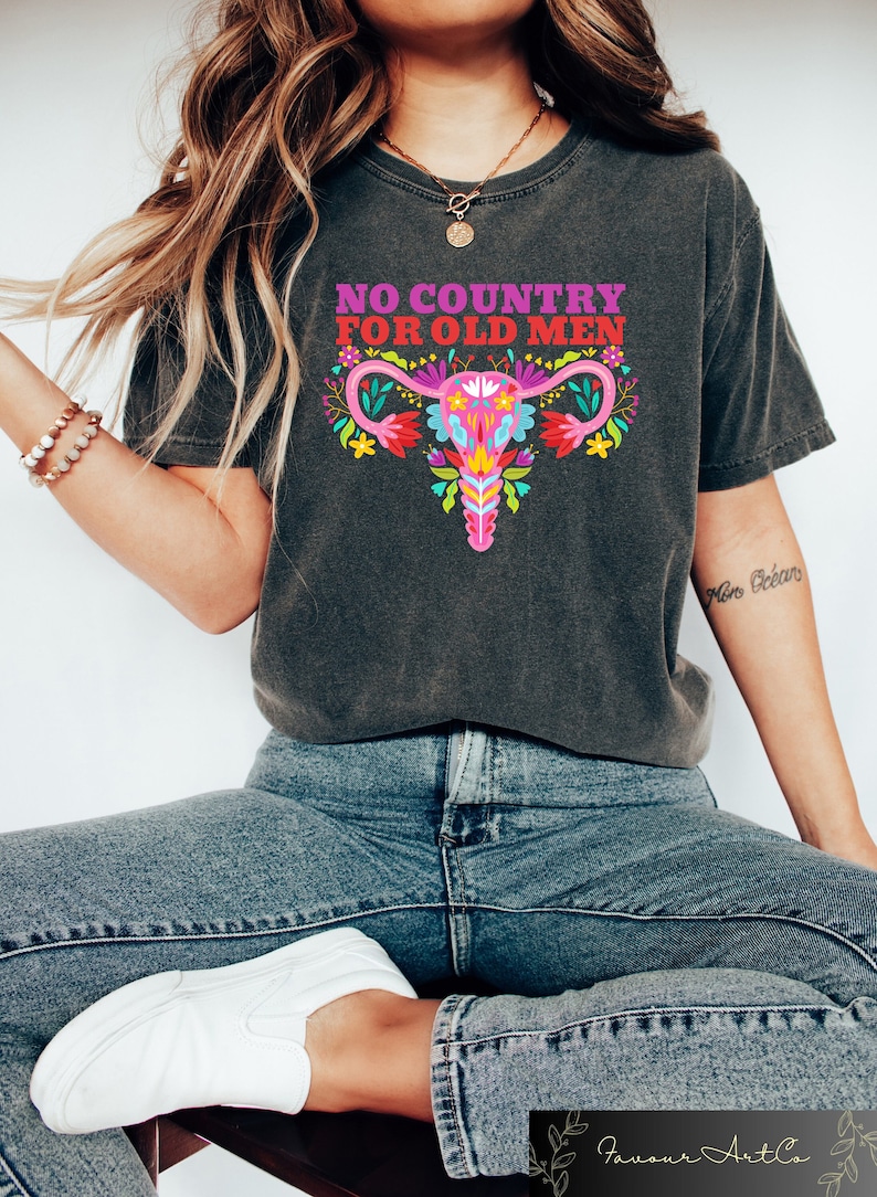 May include: A dark gray t-shirt with the text "NO COUNTRY FOR OLD MEN" and a colorful floral illustration of a uterus.