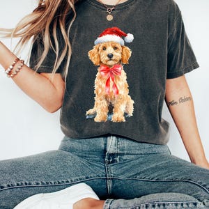 May include: Dark gray t-shirt featuring a watercolor illustration of a golden doodle puppy wearing a Santa hat and a red bow. The puppy is sitting, with a sweet expression. The shirt is a comfortable, casual style.
