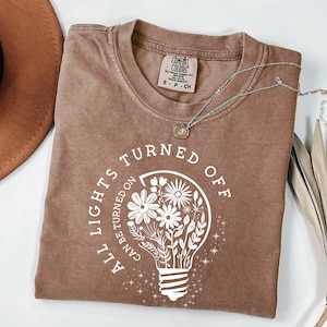 May include: A brown Comfort Colors t-shirt with the text "ALL LIGHTS TURNED OFF CAN BE TURNED ON" surrounding a lightbulb graphic filled with flowers. A silver necklace with a small pendant is on the shirt.
