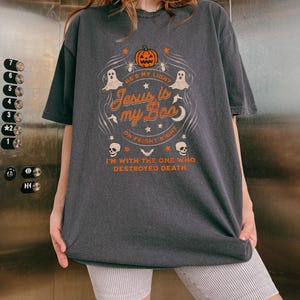 May include: Dark gray t-shirt with a Halloween-themed graphic. The design includes a pumpkin, ghosts, and the text "He's my light, Jesus is my Boo, on Fright Night, I'm with the one who destroyed death."