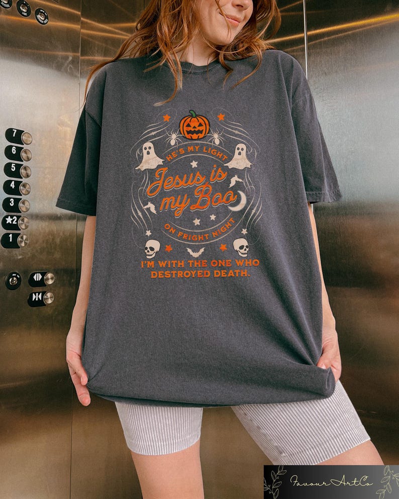 May include: Dark gray t-shirt with a Halloween-themed graphic. The design includes a pumpkin, ghosts, and the text "He's my light, Jesus is my Boo, on Fright Night, I'm with the one who destroyed death."