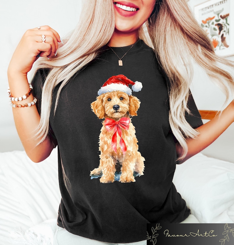 May include: Black t-shirt featuring a watercolor illustration of a golden doodle wearing a Santa hat and a red bow. The dog is sitting, with a sweet expression. The shirt has a crew neck and short sleeves. The brand name "Favour Art Co" is at the bottom.