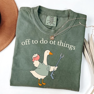 May include: An olive green short-sleeve t-shirt with a crew neckline. The shirt features a graphic of a white goose wearing a green shirt, holding a pink object and a gray reacher grabber tool. The text "off to do ot things" is printed in white.