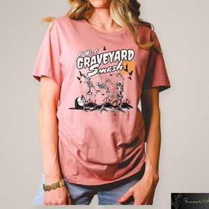 K&ouml;nnte beinhalten: Ein pinkfarbenes T-Shirt mit einem grafischen Aufdruck von Skeletten, die auf einem Friedhof tanzen. Der Text "It Was a Graveyard Smash" ist auf das Shirt gedruckt.