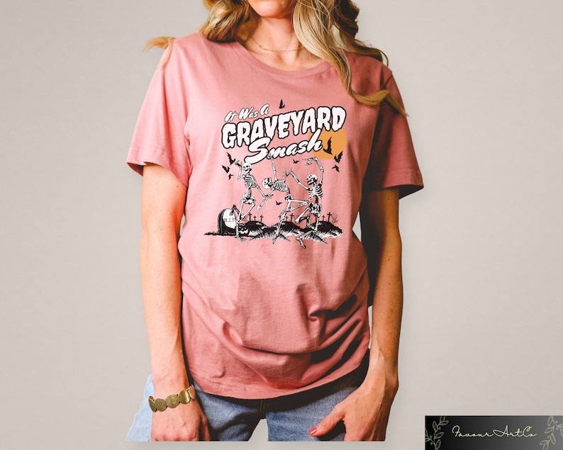 K&ouml;nnte beinhalten: Ein pinkfarbenes T-Shirt mit einem grafischen Aufdruck von Skeletten, die auf einem Friedhof tanzen. Der Text "It Was a Graveyard Smash" ist auf das Shirt gedruckt.