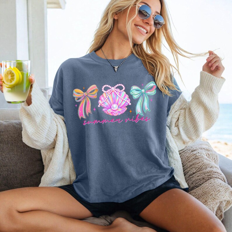 K&ouml;nnte beinhalten: Blaues T-Shirt mit einer Grafik von drei Schleifen und einer Muschel mit dem Text "summer vibes" in Pink. Die Schleifen sind in Rosa-, Gelb- und Blaut&ouml;nen gehalten. Die Muschel ist rosa und lila mit einer Perle in der Mitte.