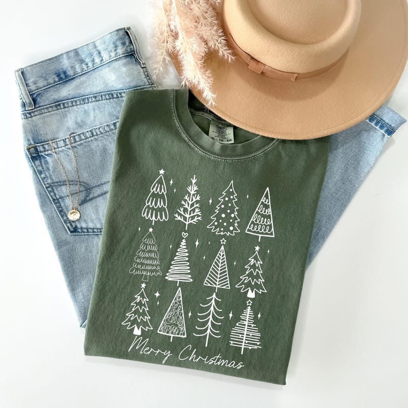 May include: An olive green t-shirt featuring a white graphic of Christmas trees and the text "Merry Christmas." The shirt is styled with light blue jeans, a gold necklace, and a tan hat with a brown band.