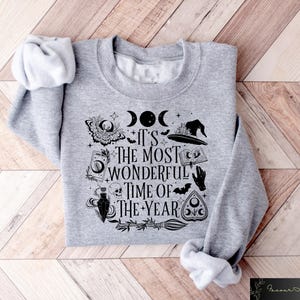 May include: Gray sweatshirt with a black and white graphic print of a witch's hat, a crescent moon, a book, a potion bottle, a skull, and a pentagram. The text on the sweatshirt reads "It's the most wonderful time of the year."
