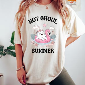 May include: Cream-colored t-shirt with the words "HOT GHOUL SUMMER" in a black, playful font. The graphic features a cartoon ghost on a pink flamingo floatie, surrounded by clouds, stars, and seashells.
