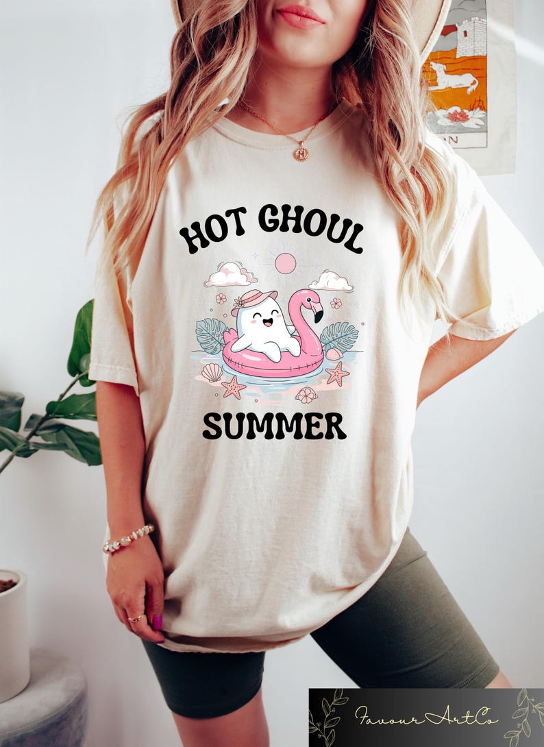 May include: Cream-colored t-shirt with the words "HOT GHOUL SUMMER" in a black, playful font. The graphic features a cartoon ghost on a pink flamingo floatie, surrounded by clouds, stars, and seashells.