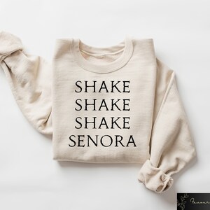 May include: A beige sweatshirt with the words "SHAKE SHAKE SHAKE SENORA" printed in black, centered on the front. The sleeves are folded, and the garment is laid flat. The sweatshirt is a casual style.