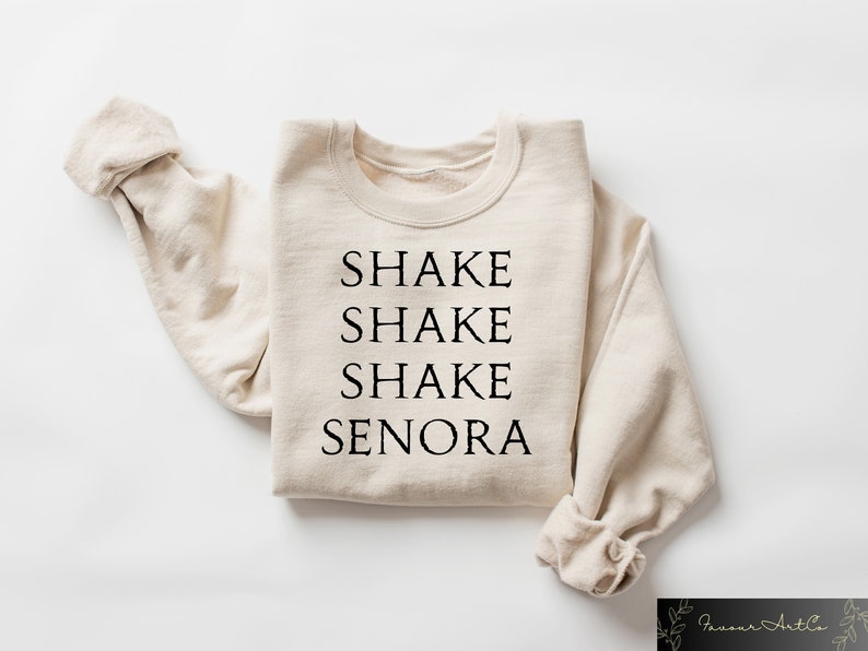 May include: A beige sweatshirt with the words "SHAKE SHAKE SHAKE SENORA" printed in black, centered on the front. The sleeves are folded, and the garment is laid flat. The sweatshirt is a casual style.