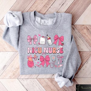 Peut inclure: Sweat-shirt gris avec un motif rose et blanc qui dit "NICU NURSE". Le motif comprend un biberon, une sucette, un st&eacute;thoscope et des n&oelig;uds.