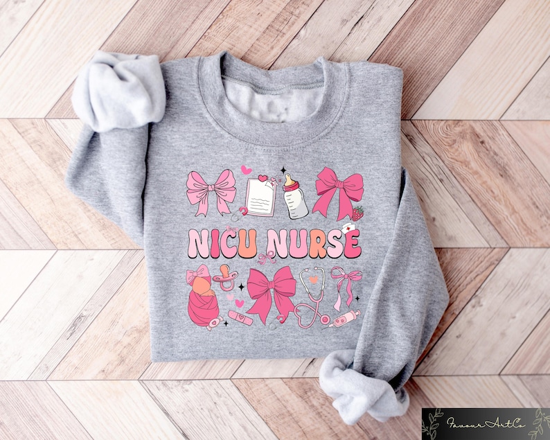Peut inclure: Sweat-shirt gris avec un motif rose et blanc qui dit "NICU NURSE". Le motif comprend un biberon, une sucette, un st&eacute;thoscope et des n&oelig;uds.