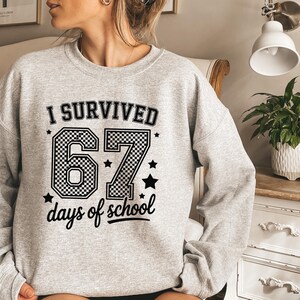 May include: A light gray sweatshirt with the text "I SURVIVED 67 days of school" in black. The number "67" is in a checkered pattern. Black stars are scattered around the text. The sweatshirt has long sleeves.