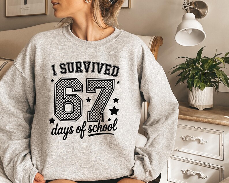 May include: A light gray sweatshirt with the text "I SURVIVED 67 days of school" in black. The number "67" is in a checkered pattern. Black stars are scattered around the text. The sweatshirt has long sleeves.