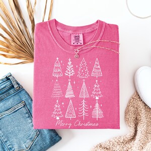 May include: A cerise t-shirt featuring a white Christmas tree design and the words "Merry Christmas." The shirt is displayed with blue jeans, white trainers, and a necklace. A dried palm leaf is also visible in the image.