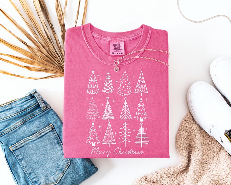May include: A cerise t-shirt featuring a white Christmas tree design and the words "Merry Christmas." The shirt is displayed with blue jeans, white trainers, and a necklace. A dried palm leaf is also visible in the image.