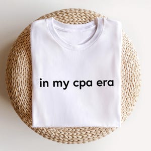 Könnte beinhalten: Ein weißes T-Shirt mit dem Aufdruck "in my cpa era" in Schwarz. Das T-Shirt ist gefaltet und liegt auf einer runden, gewebten, beigefarbenen Oberfläche. Das T-Shirt ist ein Freizeitkleidungsstück.