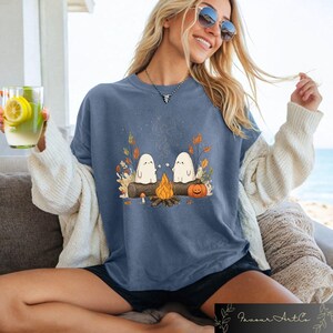 Puede incluir: Camiseta azul con una escena de fogata con dos fantasmas de dibujos animados asando malvaviscos. El dise&ntilde;o incluye una calabaza, hojas de oto&ntilde;o y un tronco. La camiseta es parte de una colecci&oacute;n con tem&aacute;tica de oto&ntilde;o o Halloween.