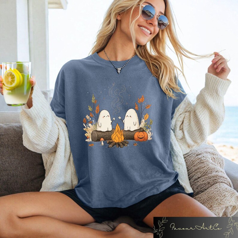 Puede incluir: Camiseta azul con una escena de fogata con dos fantasmas de dibujos animados asando malvaviscos. El dise&ntilde;o incluye una calabaza, hojas de oto&ntilde;o y un tronco. La camiseta es parte de una colecci&oacute;n con tem&aacute;tica de oto&ntilde;o o Halloween.