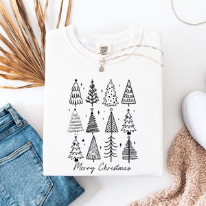 May include: White crew neck sweatshirt featuring a black graphic of Christmas trees and the words "Merry Christmas". The sweatshirt is folded and displayed with blue jeans, white shoes, and a necklace.