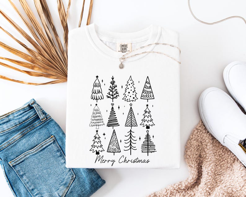 May include: White crew neck sweatshirt featuring a black graphic of Christmas trees and the words "Merry Christmas". The sweatshirt is folded and displayed with blue jeans, white shoes, and a necklace.
