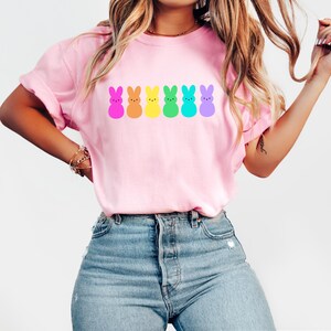 K&ouml;nnte beinhalten: Ein pinkfarbenes T-Shirt mit einer Regenbogenreihe von Cartoon-Peeps-Marshmallow-Hasen. Die Hasen sind in den Farben des Regenbogens: lila, blau, gr&uuml;n, gelb, orange und pink.