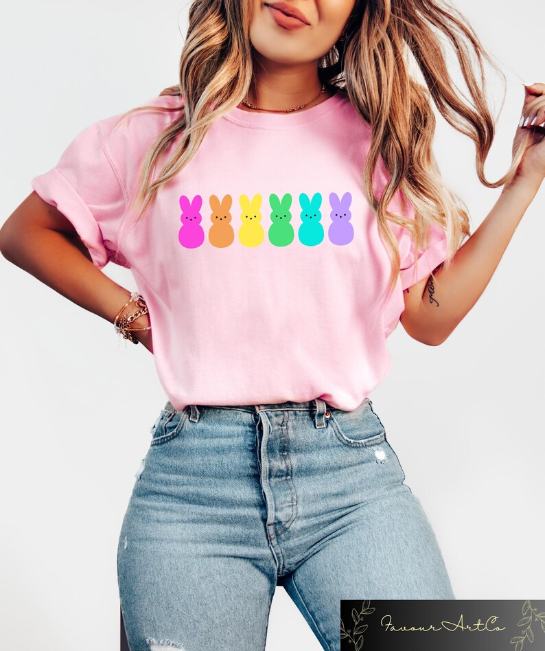 K&ouml;nnte beinhalten: Ein pinkfarbenes T-Shirt mit einer Regenbogenreihe von Cartoon-Peeps-Marshmallow-Hasen. Die Hasen sind in den Farben des Regenbogens: lila, blau, gr&uuml;n, gelb, orange und pink.