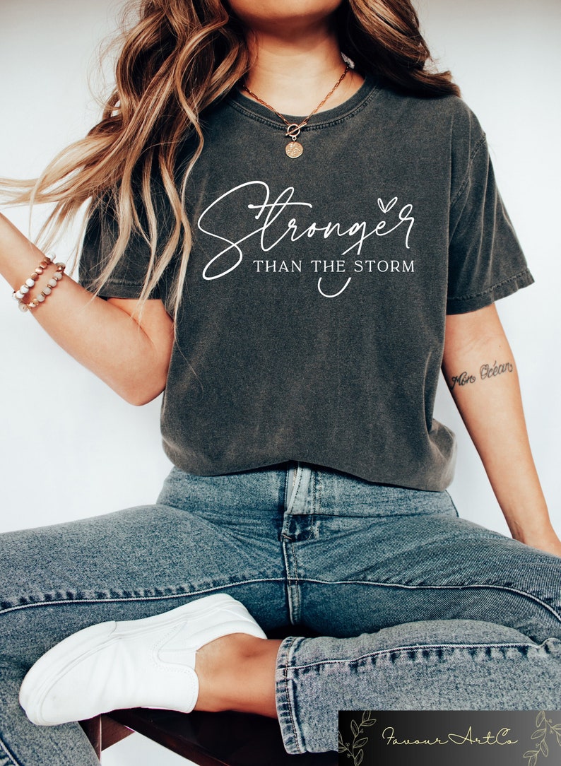 May include: A dark gray t-shirt with the text "Stronger than the storm" in white lettering. The shirt has a relaxed fit and a crew neck.