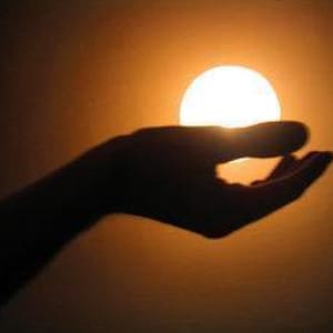 Può includere: Silhouette di una mano che tiene un globo luminoso, simile al sole. La mano è scura contro uno sfondo caldo, arancione-giallo, creando un contrasto drammatico. L'immagine evoca la sensazione di tenere o interagire con la luce.