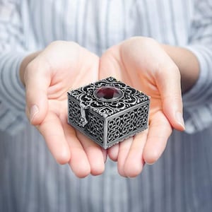 May include: A square, silver-toned metal trinket box featuring intricate, openwork filigree patterns on all sides. The lid is adorned with a large, faceted, deep red gemstone set in the center, surrounded by detailed metal scrollwork.